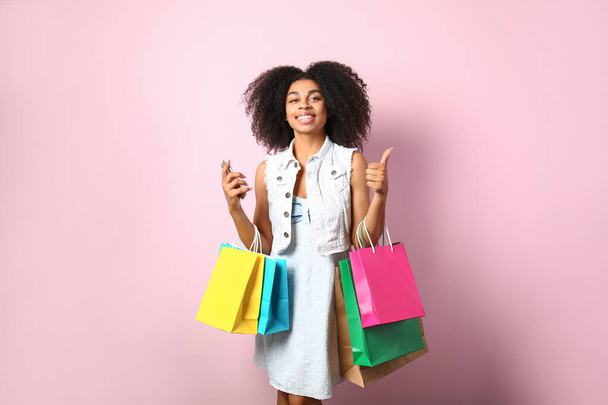 Beautiful African-American woman with shopping bags and mobile phone showing thumb-up on color background - Photo, Image