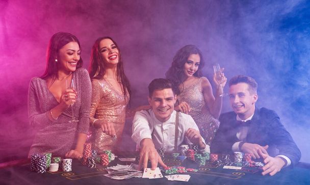 Ystäviä pelaamassa pokeria kasinolla. He juhlivat voittoaan, juovat samppanjaa ja poseeraavat pöydässä sipsien, rahan ja korttien kanssa.
. - Valokuva, kuva