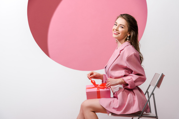 happy woman sitting on chair and holding present on white and pink  - 写真・画像