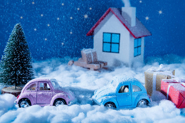 Small blue pink toy cars stand outdoors on snowy weather with red yellow giftboxes green pine tree white joyful family celebrate christmas eve in white comfort house over blue sky background - Zdjęcie, obraz