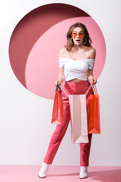 έκπληκτη κοπέλα με γυαλιά ηλίου κρατώντας τσάντες για ψώνια σε λευκό και ροζ  - Φωτογραφία, εικόνα