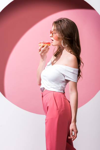 side view of beautiful girl holding tasty doughnut and standing on pink and white  - Fotografie, Obrázek