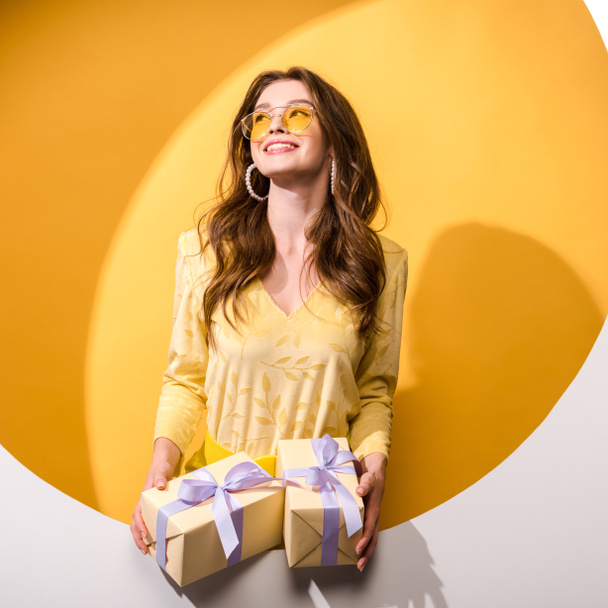 Ευτυχισμένη γυναίκα με γυαλιά ηλίου κρατώντας δώρα σε πορτοκαλί και άσπρο  - Φωτογραφία, εικόνα
