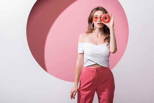 atractiva chica cubriendo la cara con sabroso donut y de pie en rosa y blanco
  - Foto, Imagen