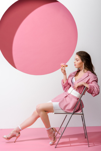 attractive woman sitting on chair and holding sweet doughnut on pink and white  - Фото, зображення