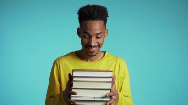 African student on blue background in the studio holds stack of university books from library. Guy smiles, he is happy to graduate. - Video