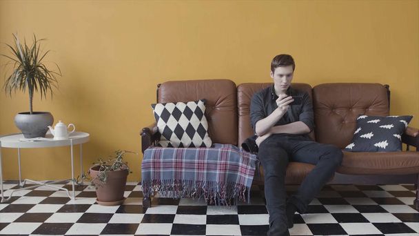 Νεαρός άνδρας που χρησιμοποιεί το κινητό τηλέφωνο για να στείλει μήνυμα ενώ κάθεται σε ένα δερμάτινο καναπέ στο δωμάτιο με μαύρο και άσπρο πάτωμα και κίτρινο τοίχο. Πλάνα μετοχών. Σύγχρονες τεχνολογίες. - Φωτογραφία, εικόνα