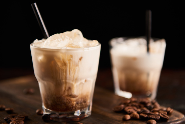 selective focus of white russian cocktail in glasses with straws on wooden board with coffee grains isolated on black - Foto, Bild