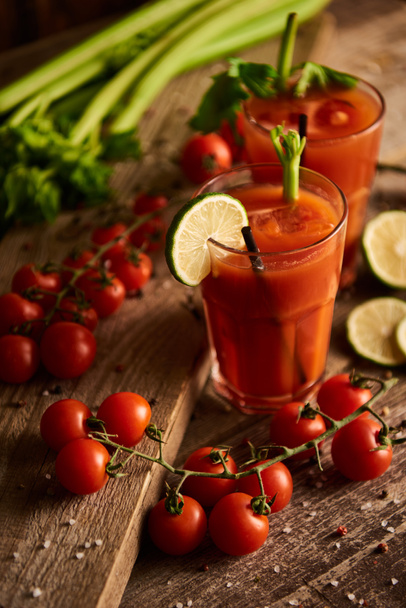  selective focus of bloody mary cocktail in glasses on wooden background with salt, pepper, lime, tomatoes and celery - Photo, Image