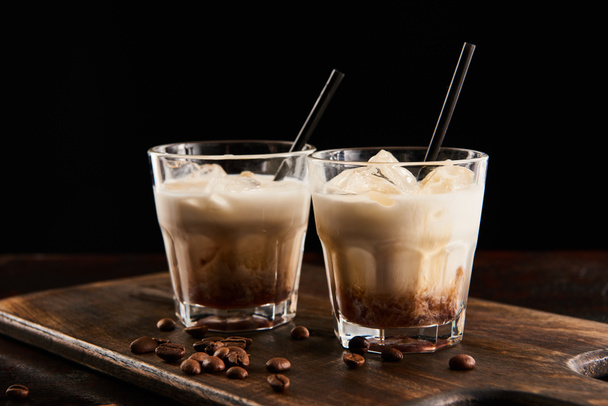 white russian cocktail in glasses with straws on wooden board with coffee grains isolated on black - Photo, image