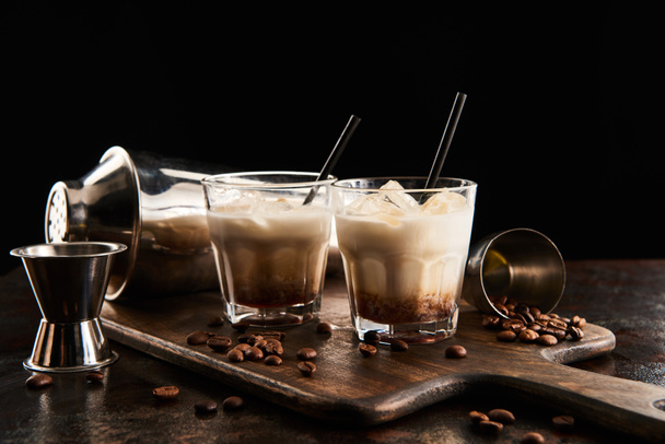 white russian cocktail in glasses with straws on wooden board with coffee grains and shaker isolated on black - Fotó, kép