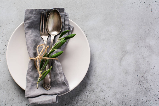 Rustic vintage set of cutlery. Plate with grey linen napkin, fork and spoon, olive tree branch over rustic concrete gray old background. Fall holiday table decoration setting. Top view, copy space. - Zdjęcie, obraz