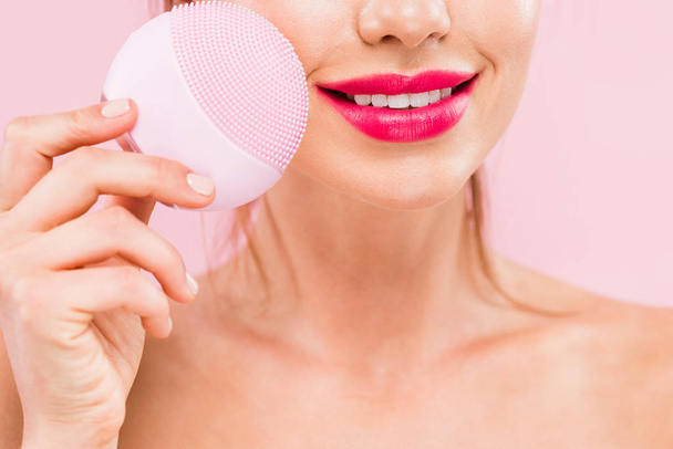 cropped view of naked beautiful woman with pink lips holding facial cleansing brush isolated on pink  - Foto, Imagen