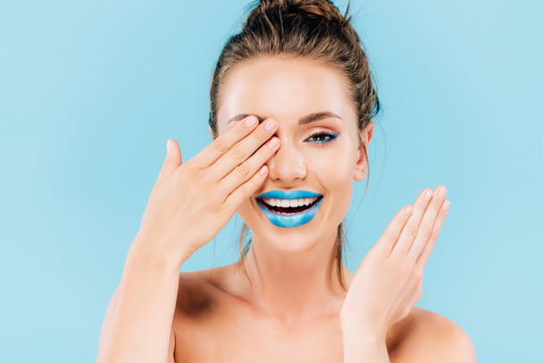 szczęśliwa piękna kobieta z niebieskimi ustami okrywająca oko dłonią odizolowaną na niebiesko - Zdjęcie, obraz