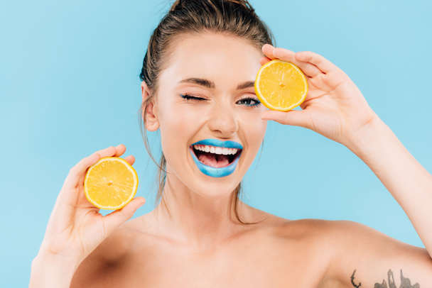 szczęśliwy mrugający nagi nagi piękny kobieta z niebieski usta trzymając pomarańczowy połówki odizolowany na niebieski - Zdjęcie, obraz