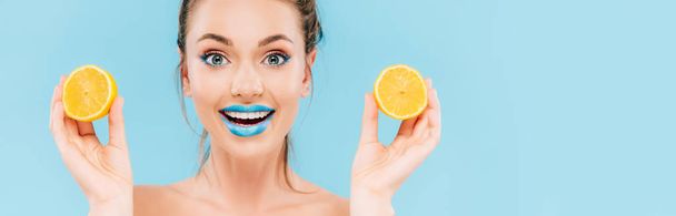 podniecona naga piękna kobieta z niebieskimi ustami trzymająca pomarańczowe połówki na niebieskim, panoramicznym ujęciu - Zdjęcie, obraz