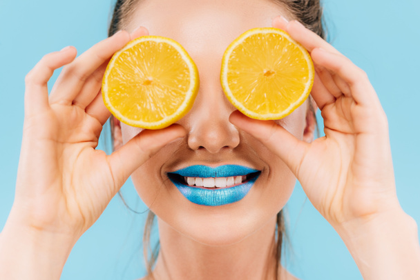 smiling beautiful woman with blue lips holding orange halves on eyes isolated on blue - Fotoğraf, Görsel