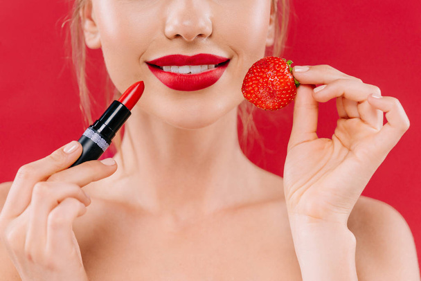 przycięty widok naga piękna kobieta z czerwonymi ustami trzymając szminkę i truskawki izolowane na czerwono - Zdjęcie, obraz