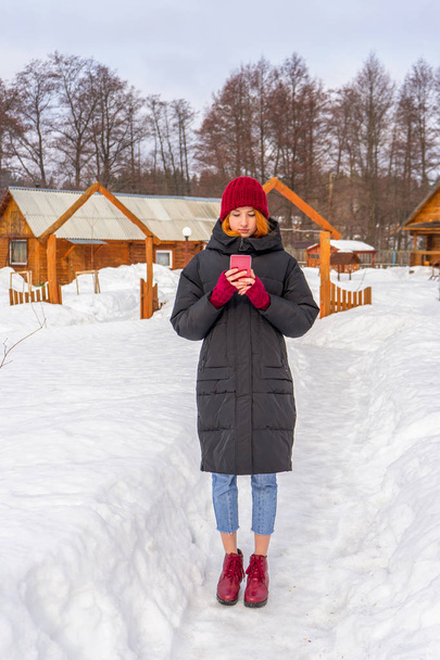 Teen cute girl in black down jacket, blue cropped jeans, burgundy hat, boots and fingerless gloves standing outdoor against winter rural landscape. Weekend at countryside, teenage fashion concept - Photo, image