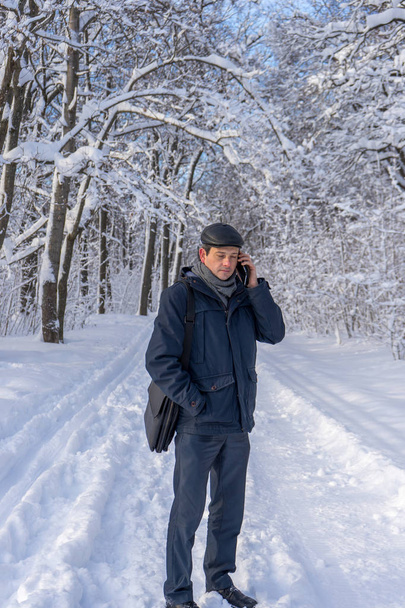 Handsome middle-aged man walking in winter snowy park or forest. Attractive man in jacket, scarf and cap talking on mobile phone. Winter mood, authentic lifestyle concept, stylish male outfit - Foto, Imagem