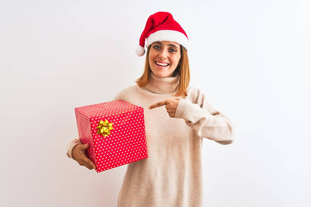 Beautiful redhead woman wearing christmas hat holding present over isolated background very happy pointing with hand and finger - Foto, imagen