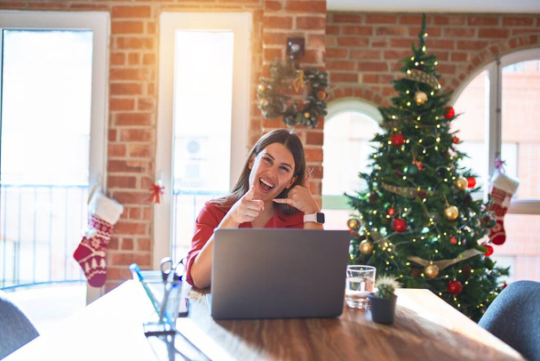 クリスマスツリーの周りの自宅でラップトップで作業テーブルに座っている美しい女性は、電話のジェスチャーで話しているとあなたを指して笑っています。電話してくれ. - 写真・画像