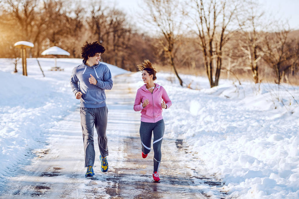 Счастливая, улыбающаяся кавказская пара в спортивной одежде, бегающая на природе, глядя друг на друга. Зимнее время
. - Фото, изображение
