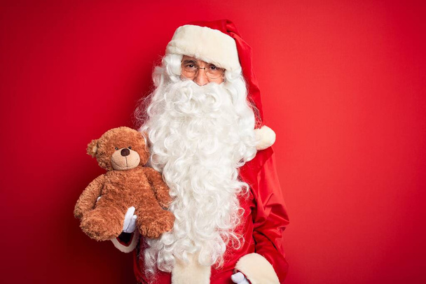 Чоловік середнього віку, одягнений в костюм Санта Клауса, тримає Тедді ведмедя над ізольованим червоним фоном з щасливим обличчям стоячи і посміхаючись з упевненою посмішкою, що показує зуби. - Фото, зображення