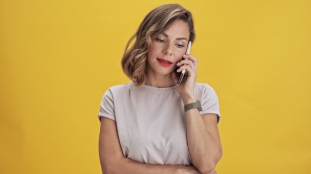 Beautiful young woman with red lips talking on mobile phone and smiling while standing over yellow background isolated - Video