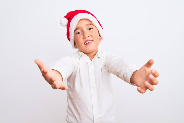 Hermoso niño con sombrero de Santa Navidad de pie sobre fondo blanco aislado mirando a la cámara sonriendo con los brazos abiertos para el abrazo. Expresión alegre abrazando la felicidad
. - Foto, imagen