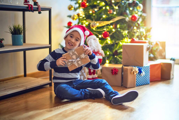 Очаровательный малыш улыбается счастливым и уверенным. Сидя на полу в шляпе Санта Клауса с улыбкой на лице, держа подарок вокруг рождественской елки дома
 - Фото, изображение