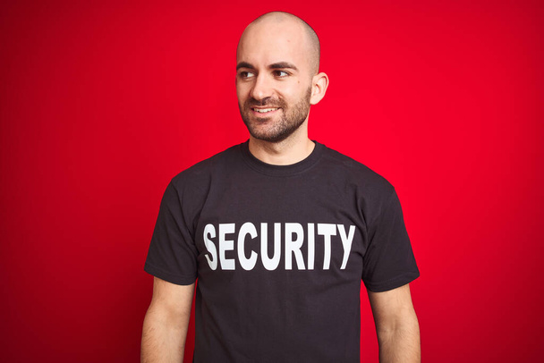 Young safeguard man wearing security uniform over red isolated background looking away to side with smile on face, natural expression. Laughing confident. - Photo, Image