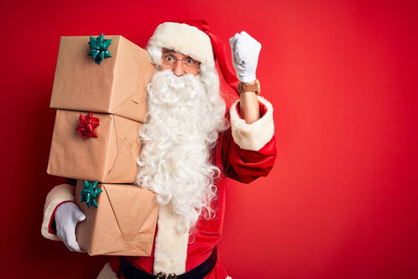 Ανώτερος άνθρωπος φορώντας κοστούμι Άγιος Βασίλης κρατώντας πύργο των δώρων πάνω από απομονωμένο κόκκινο φόντο ενοχλημένος και απογοητευμένος φωνάζοντας με θυμό, τρελός και φωνάζοντας με σηκωμένο χέρι, έννοια του θυμού - Φωτογραφία, εικόνα