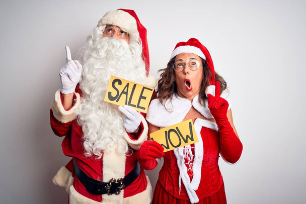Pari yllään Santa puku tilalla wow ja myynti banneri yli eristetty valkoinen tausta hämmästynyt ja yllättynyt katselee ylös ja osoittaa sormilla ja nosti kädet
. - Valokuva, kuva