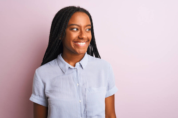 縞模様のシャツを着た若いアフリカ系アメリカ人女性がピンクの背景に立って顔に笑顔で横に離れて見て、自然な表情。自信を持って. - 写真・画像