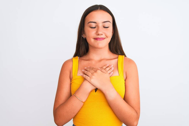 Young beautiful girl wearing yellow casual t-shirt standing over isolated white background smiling with hands on chest with closed eyes and grateful gesture on face. Health concept. - Foto, imagen
