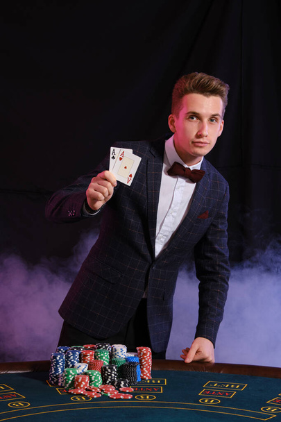 Елегантний чоловік грає в покер в казино. Він показує два аси під час позування за столом зі стопками чіпсів на ньому, на чорному тлі
. - Фото, зображення