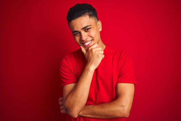 Jeune homme brésilien portant t-shirt debout sur fond rouge isolé regardant avec confiance la caméra souriant avec les bras croisés et la main levée sur le menton. Penser positif
. - Photo, image