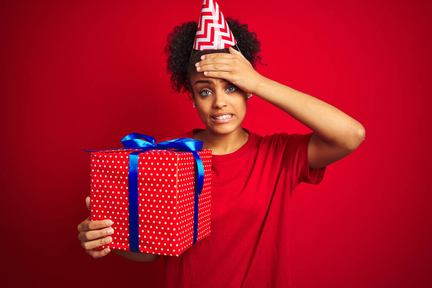 Komik şapkalı Afrikalı bir kadın izole edilmiş kırmızı arka planda doğum günü hediyesi tutuyor. Elleri başının üstünde, utanç ve şaşkınlık dolu, sinirli ve hayal kırıklığına uğramış bir halde. Hata için korku ve üzüntü. - Fotoğraf, Görsel