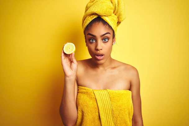 Afro femme portant serviette après la douche tenant tranche de citron sur fond jaune isolé peur en état de choc avec un visage surprise, peur et excité par l'expression de la peur
 - Photo, image