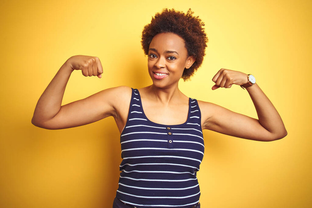 孤立した黄色の背景の上に夏のTシャツを着て、誇らしげに腕の筋肉を示すボートゥールアフリカ系アメリカ人女性。フィットネスコンセプト. - 写真・画像