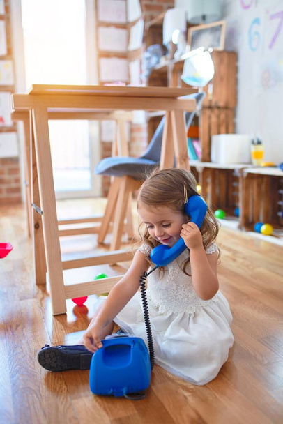 Adorável criança loira brincando com telefone vintage. Sentado no chão em torno de lotes de brinquedos no jardim de infância
 - Foto, Imagem