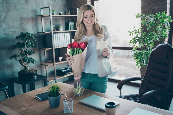 Photo of beautiful blond business lady came workplace morning holding little postcard read corporate party invitation fresh tulips bunch table modern cozy office - Fotó, kép