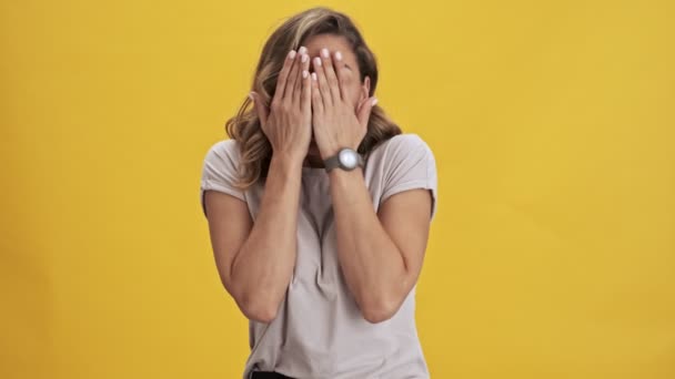 Frighten and scared young woman with red lips covers her face with her hands and peeping through her fingers over yellow background isolated - Filmagem, Vídeo