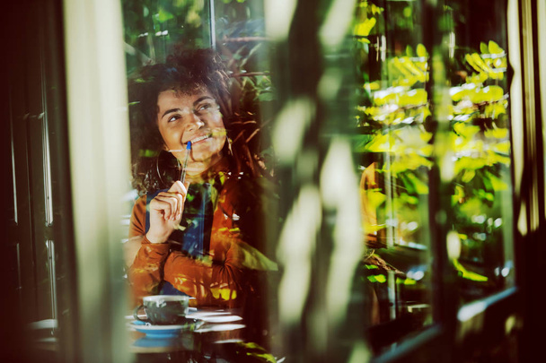 Upea hymyilevä sekoitettu rotu nuori nainen kihara tukka istuu kahvilassa, pitää päiväkirjaa rinnassa ja katselee pois. Ulkoa otettu kuva
. - Valokuva, kuva