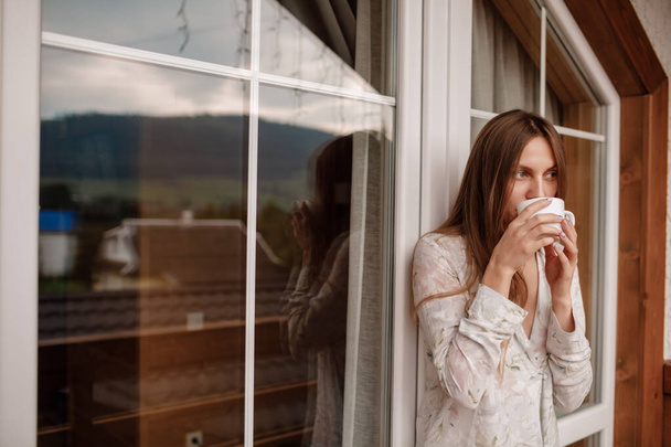 Portrait of a happy smiling young woman in stylish nightwear with a cup of coffee or tea meeting the day standing on the hotel terrace with a gorgeous view of the mountains. place for inscription - 写真・画像