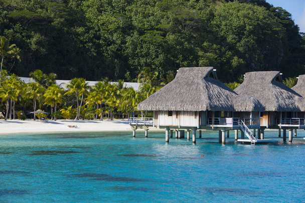 Widok z niebieskiego tropikalnego morza autentycznych tradycyjnych domów polinezyjskich z krytym strzechą dachem nad wodą i piaszczystej plaży z palmami i góry w tle. Polinezja, Tahiti - Zdjęcie, obraz