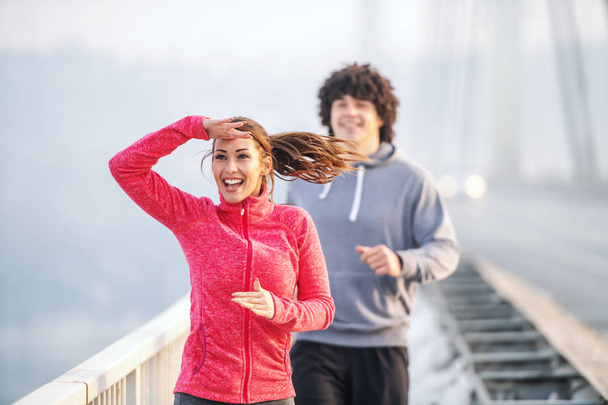 Glimlachend gelukkig Kaukasisch echtpaar in sportkleding lopend op brug.Vrouw gespot vriend. Wintertijd. Outdoor fitness concept. - Foto, afbeelding