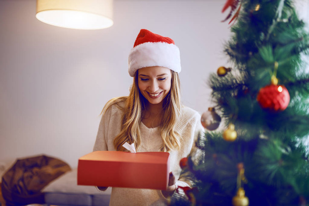 Belle femme blonde caucasienne avec chapeau de Père Noël sur la tête souriant et tenant cadeau de Noël tout en se tenant debout dans le salon. Au premier plan est décoré sapin
. - Photo, image