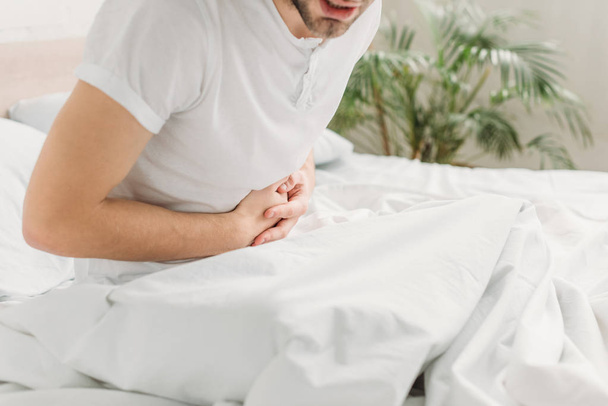 cropped view of man sitting in bed and suffering from abdominal pain - Foto, imagen